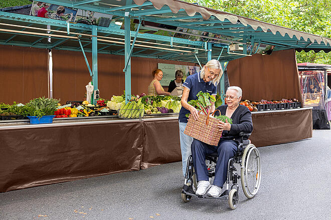Nachbarschaftshilfekurs im Oktober Eine Johanniterin reicht einer Frau, die in einem Rollstuhl sitzt, einen Korb gefüllt mit Gemüse. Sie befinden sich vor einem Marktstand.