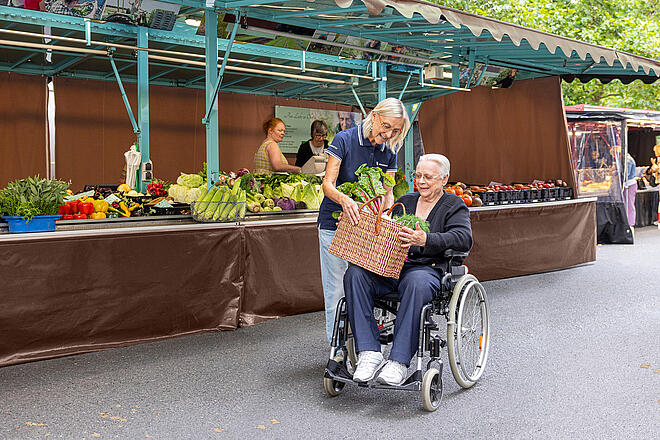 Nachbarschaftshilfekurse Eine Frau reicht einer älteren Frau, die in einem Rollstuhl sitzt, einen Korb mit Gemüse. Sie befinden sich auf einem Wochenmarkt.