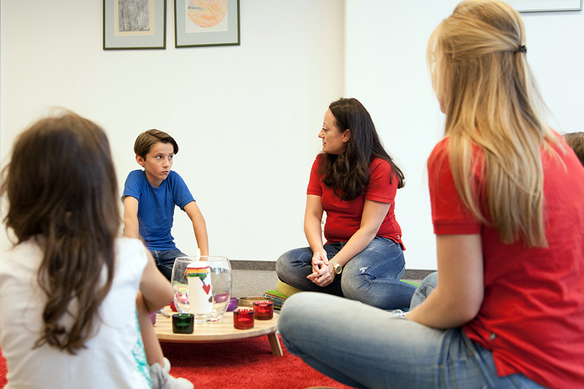 Kindergruppe im Gespräch mit Betreuerin in einem Raum mit Kerzen und Bildern an der Wand.
