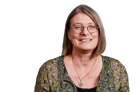 Frau mit Brille und mittellangen Haaren lächelt in die Kamera, trägt gemusterte Bluse und Halskette.