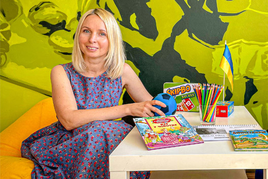 Eine junge Frau sitzt an einem Tisch mit Kinderspielen, bunten Stiften und Kinderbüchern. Außerdem steht auf dem Tisch eine kleine Ukraine-Flagge.