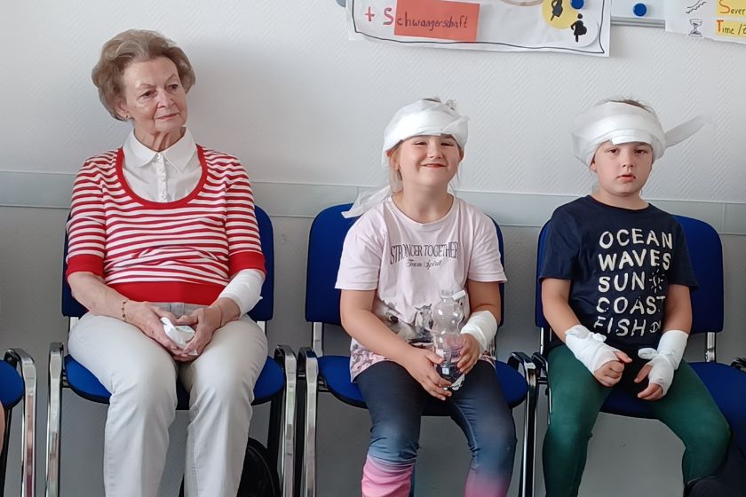 Eine ältere Dame mit Verband am Arm sitzt auf einem Stuhl neben einem Kind mit Verband am Kopf. Die Dame schaut nachdenklich, das Kind lacht fröhlich.