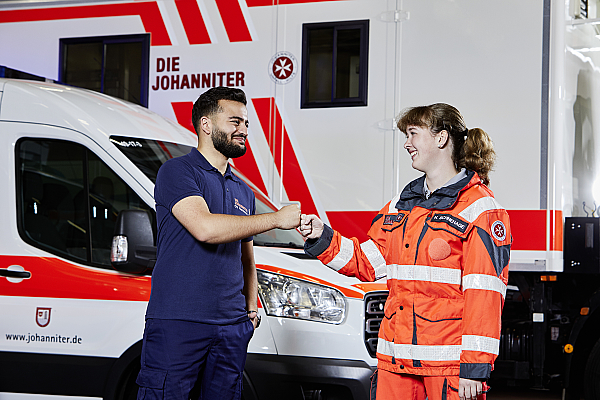 Ein BFDler und eine BFDlerin der Johanniter begrüßen sich, im Hintergrund sind Fahrzeuge der Johanniter.