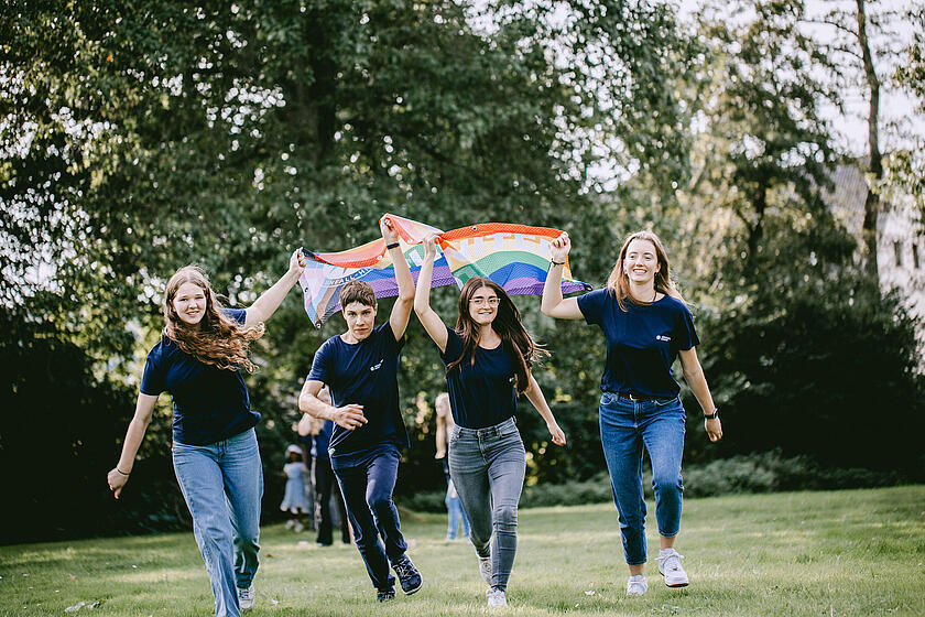 Mehrere Jugendliche rennen mit einer Regenbogenflagge über eine Wiese