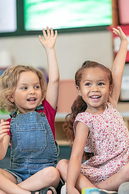 Eine Gruppe Vorschulkinder sitzt im Schneidersitz auf dem Boden und zeigt mit den Händen auf, um die Frage ihres Lehrers zu beantworten.