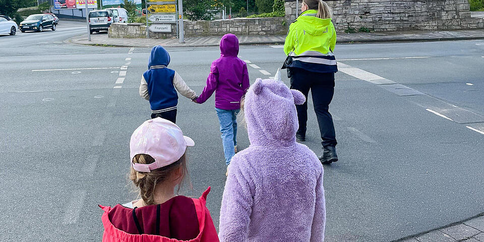 Kinder gehen in Zweipaaren Hand in Hand mit einer Polizistin über einen Fußgängerüberweg
