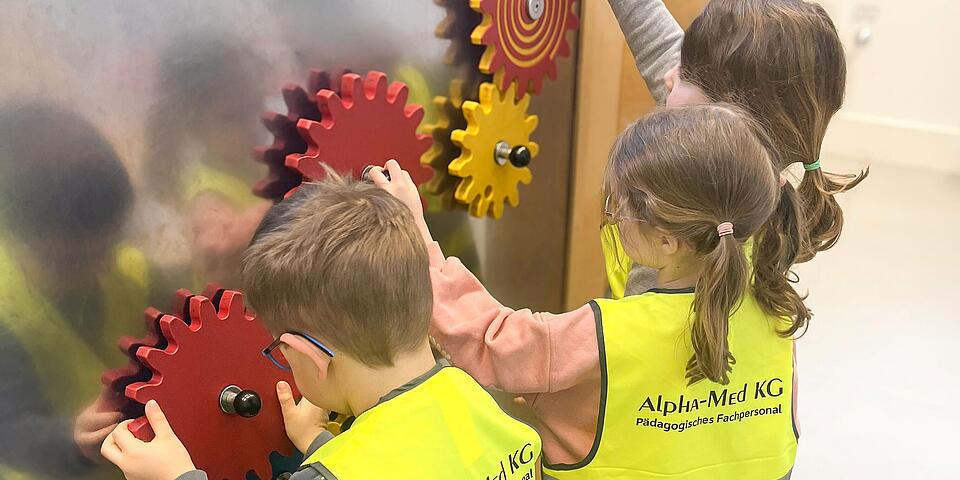 Drei Kinder mit gelben Warnwesten an einer Metallwand an der Zahnräder zum Drehen sind.