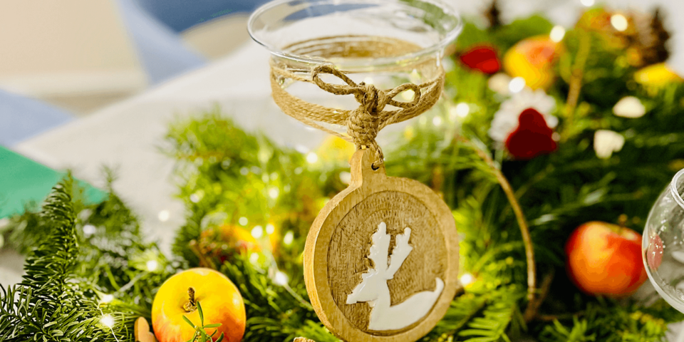Nahaufnahme einer festlichen Tischdeko mit Tannenzweigen, gläsernem Windlicht, Holzanhänger mit Rentier-Motiv und Äpfeln.
