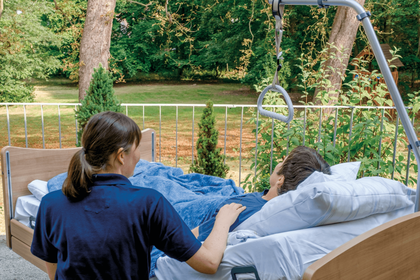 Ein Mensch mit kurzen dunklen Haaren liegt auf einer Terrasse in einem Bett und sieht in den Garten. Neben sitzt eine Frau in einem blauen Poloshirt und berührt seine Schulter.