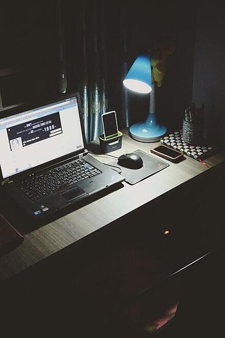 Ein beleuchteter Schreibtisch in einem dunklen Raum mit Laptop, Schreibtischlampe, Smartphone, Notizbuch und Lichterkette im Hintergrund.