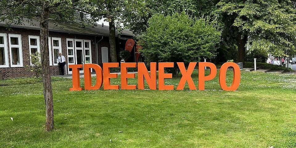 Orangene Großbuchstaben auf einer Wiese. Zu lesen ist "Ideenexpo"