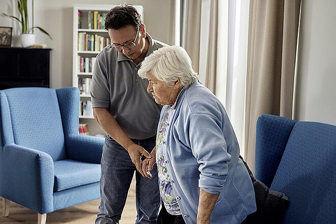Pflegeszene zu Hause: Ein Mann gibt einer Seniorin Halt beim sicheren Aufstehen.