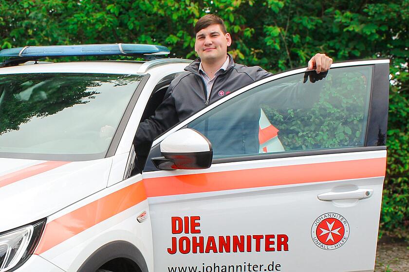 Ein junger Mann mit Johanniter-Jacke steht an einem Johanniter-Fahrzeug und lehnt an der geöffneten Tür.