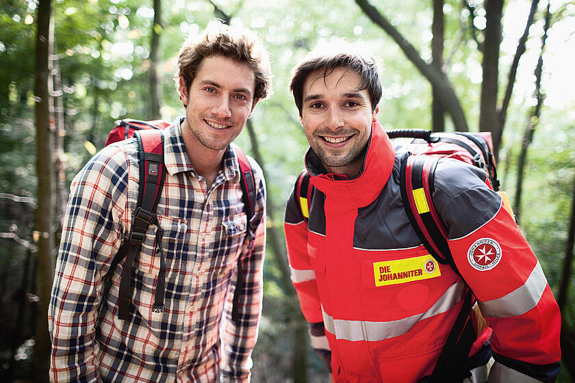 Ein Johanniter und ein weiterer Mann stehen nebeneinander in einem Wald und lächeln in die Kamera.