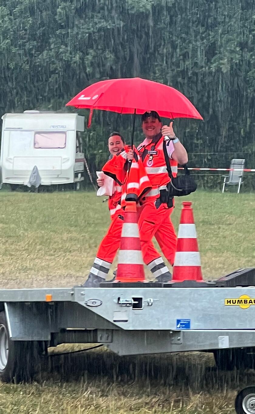 Zwei Einsatzkräfte der Johanniter in leuchtender Einsatzkleidung gehen bei starkem Regen über eine Wiese. Sie halten einen großen roten Regenschirm und lächeln in die Kamera, während eine Person den Daumen nach oben zeigt. Im Hintergrund sind ein Wohnwagen und grüne Bäume zu sehen. Die Szene vermittelt Teamgeist und gute Laune trotz widriger Wetterbedingungen beim Sanitätsdienst auf dem Parookaville Festival.