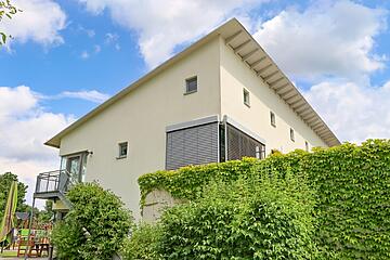 Außenansicht eines hellen zweistöckigen Gebäudes mit Schrägdach, umgeben von viel Grün und blauem Himmel.