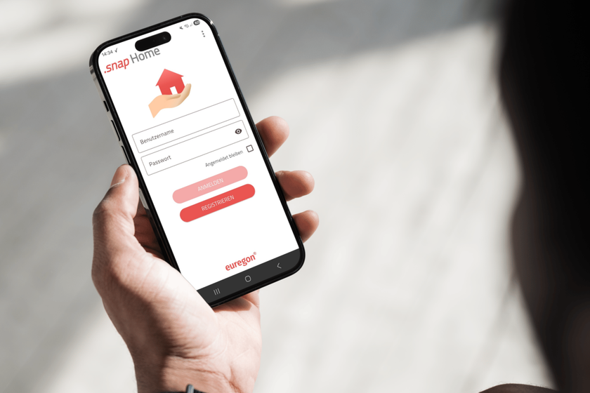 Hand hält Smartphone mit Login‑Maske zur snapHome-App; Felder für Benutzername/Passwort und rote Buttons sichtbar