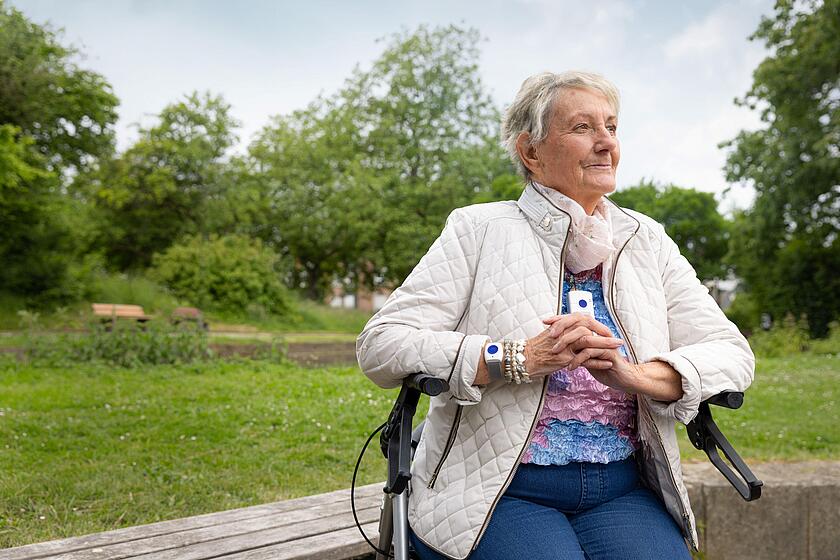 Eine Seniorin in weißer Steppjacke sitzt in einem Park auf einem Rollator. Als Halskette sowie um das Handgelenk trägt sie jeweils ein weißes rechteckiges Gerät mit einem blauen Knopft