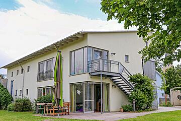 Zweistöckiges, modernes helles Gebäude mit Außentreppe, großen Fenstern und Grünfläche im Vordergrund.