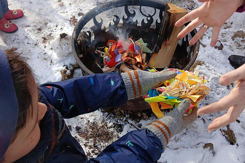 Kinder sitzen vor einem Lagerfeuer und werfen Faschingsluftschlangen in die Glut. Außenrum ist es weiß, denn es hat geschneit.