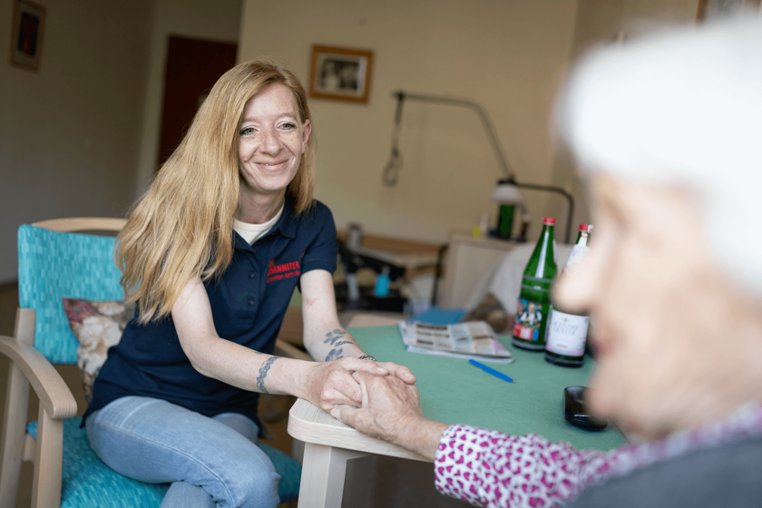 Pflegerin hält Hand einer älteren Frau am Tisch, freundliche Atmosphäre im Pflegeheim.