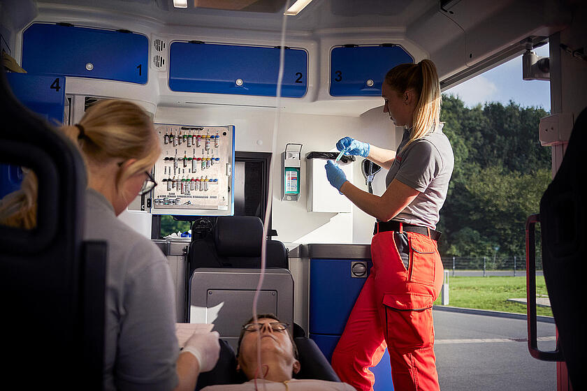 Sanitäterinnen der Johanniter versorgen einen Patienten im Rettungswagen. Eine Sanitäterin bereitet Medikamente vor, während die andere am Kopf des Patienten sitzt und Unterlagen prüft.