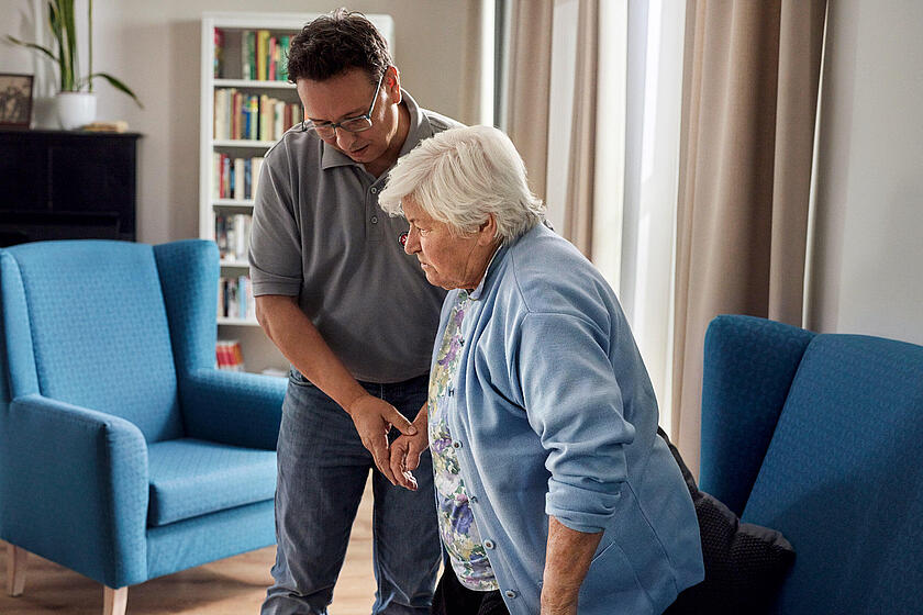 Ein Mitarbeiter der Johanniter stützt eine Seniorin beim Aufstehen aus einem Sessel.