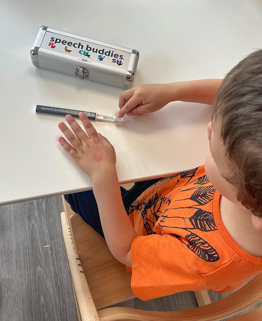 Ein Kind mit orangefarbenem T-Shirt sitzt an einem Tisch und hält ein Logopädie-Hilfsmittel namens „speech buddies“ in der Hand. Auf dem Tisch liegt ein silbernes Etui mit der Aufschrift „speech buddies“ und bunten Symbolen.