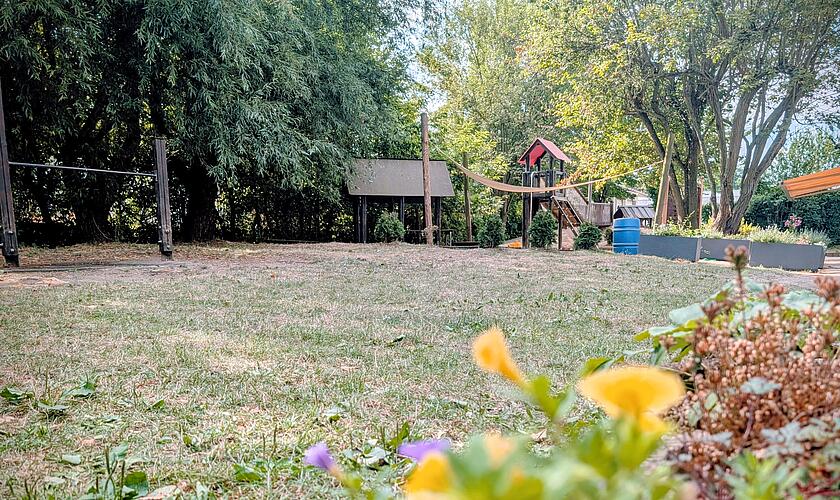 Blick über das Außengelände der Kita Erkrath: Im Vordergrund Blumen, dahinter Wiese, Sandspielbereich und Spielhäuschen
