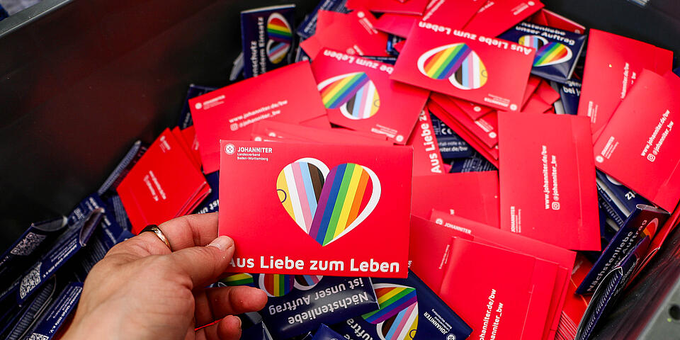 Eine Person hält eine Postkarte der Johanniter in Rot, mit einem Regenbogenherz und der Aufschrift "Aus Liebe zum Leben" und dem Johanniter-Logo. Im Hintergrund ist eine Kiste mit vieler dieser Karten, auch in blau.