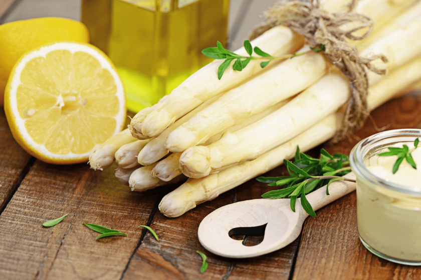 Ein Bündel weißer Spargel liegt zwischen einer aufgeschnittenen Zitrone und einem hölzernen Kochlöffel, auf dem ein kleines Herz zu sehen ist