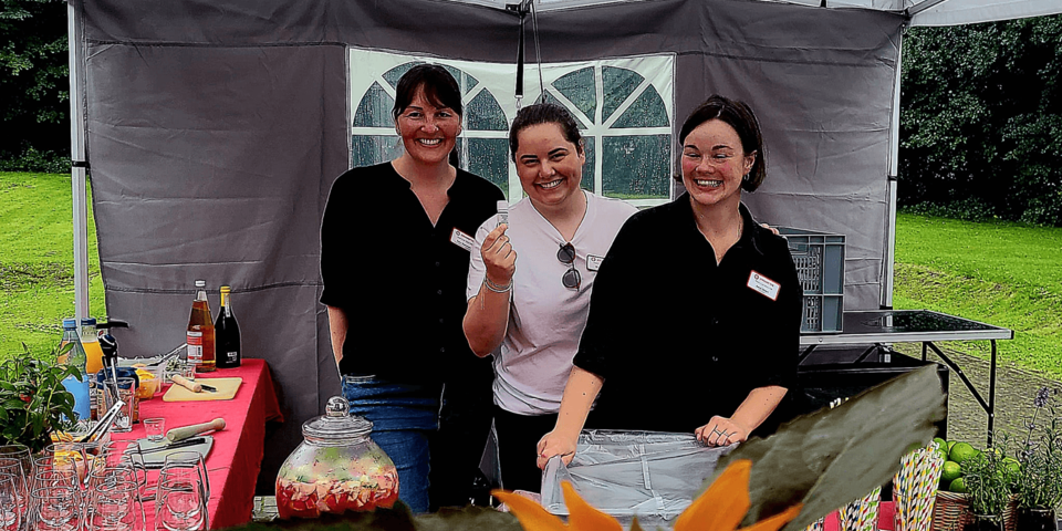 drei lächelnde junge Frauen stehen unter einem halboffenen Pavillon. Vor ihnen sind Getränke und Gläser