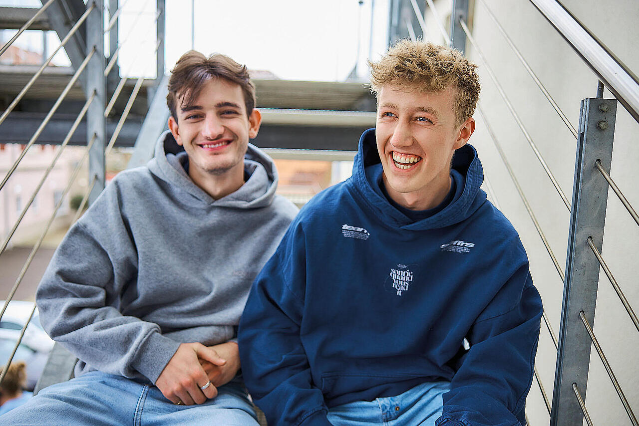 zwei junge Männer sitzen auf einer Metalltreppe und lächeln in die Kamera