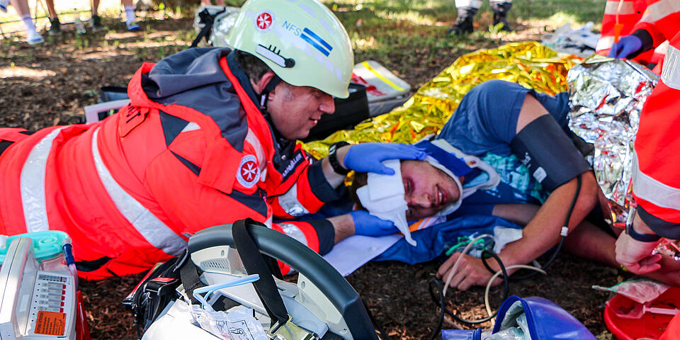 Rettungshelfer*innen stabilisieren im Freien einen verletzten Mann, medizinische Geräte und Notfallausrüstung liegen im Vordergrund.