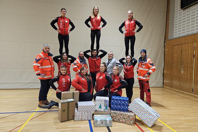 Stadtgarde und Einsatzkräfte in roten Jacken bildet Menschenpyramide in Turnhalle, Geschenke vorne.
