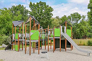 Großes, modernes Klettergerüst auf einem Spielplatz mit grünen und grauen Elementen, mehreren Plattformen, Kletterstangen und einer Rutsche; im Hintergrund Bäume und Grün.