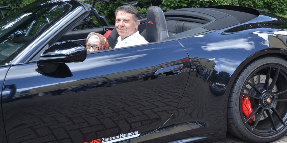 Eine weißhaarige Frau und ein größere Mann sitzen nebeneinander in einem schwarzen Auto mit offenem Verdeck.