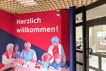 Ein großes Wandbild mit Illustrationen von Menschen verschiedenen Alters und der Aufschrift "Herzlich willkommen" begrüßt die Gäste im Begegnungsraum Liederbach.