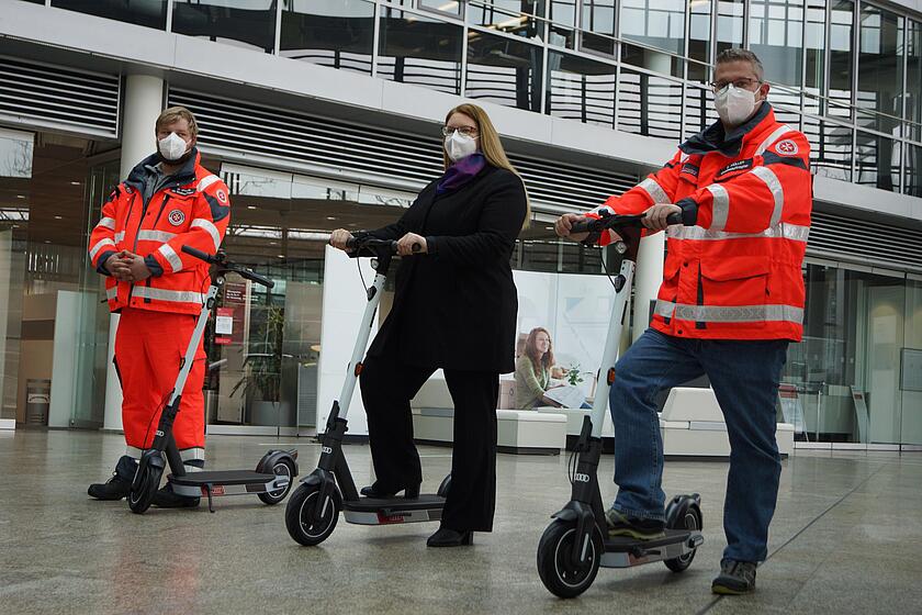Zwei Rettungskräfte in roter Einsatzkleidung und eine Frau in schwarzer Kleidung stehen mit E-Scootern in einer modernen Eingangshalle und tragen FFP2-Schutzmasken.