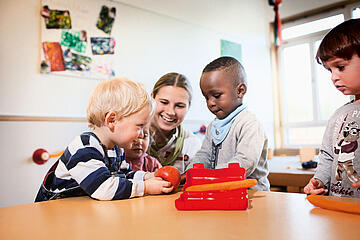 Erzieherin sitzt mit vier Kindern an einem Tisch beim gemeinsamen Basteln und Spielen.