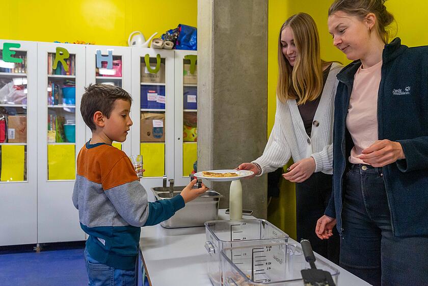 Ein Junge übergibt seinen Teller mit Essensresten zwei Studentinnen.