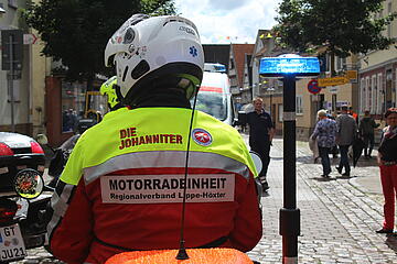 Ein Mitglied der Johanniter-Motorradstaffel Lippe-Höxter sichert eine städtische Veranstaltung ab. Im Hintergrund laufen weitere Personen. 