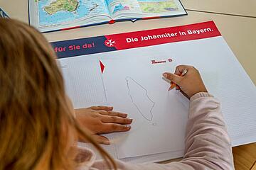Kind zeichnet auf einem Arbeitsblatt mit der Überschrift „Die Johanniter in Bayern“, daneben ein aufgeschlagener Atlas