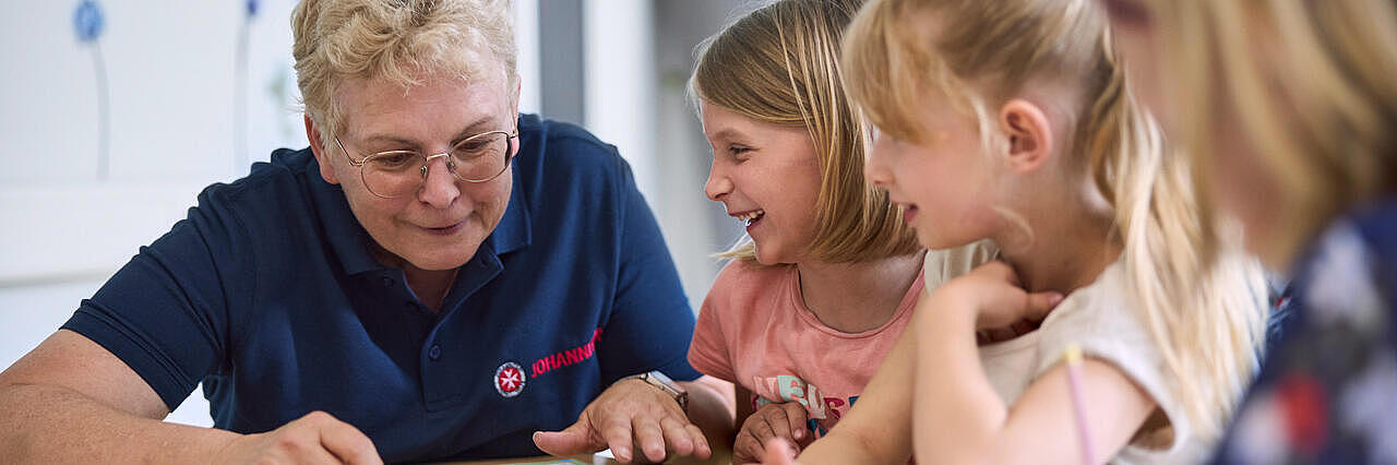 Johanniter-Mitarbeiterin liest mit Kindern ein Erste-Hilfe-Heft in fröhlicher Runde