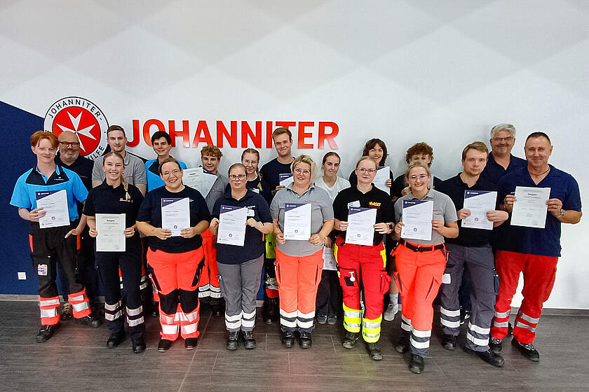 17 junge Menschen mit Zertifikaten in den Händen und in Einsatzbekleidung und zwei Männer mit blauen Johanniter-Polo-Shirts. Im Hintergrund eine Wand mit Johanniter-Logo.