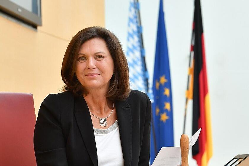 Frau sitzt mit Dokumenten am Rednerpult, hinter ihr Flaggen von Deutschland, Bayern und der EU.