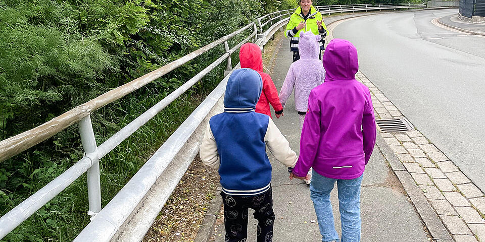 4 Kinder folgen Hand in Hand in 2 Paaren einer Polizistin auf einem Fußweg neben einer Straße