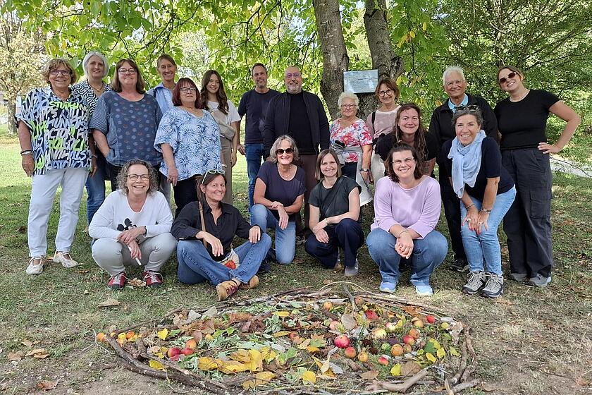 Gruppenfoto der Johanniter-Mitarbeitenden beim Lacrima-Mitarbeitendenwochenende unter Bäumen im Freien. Davor liegt ein herbstliches Mandala aus Ästen, Blättern, Tannenzapfen und Äpfeln.