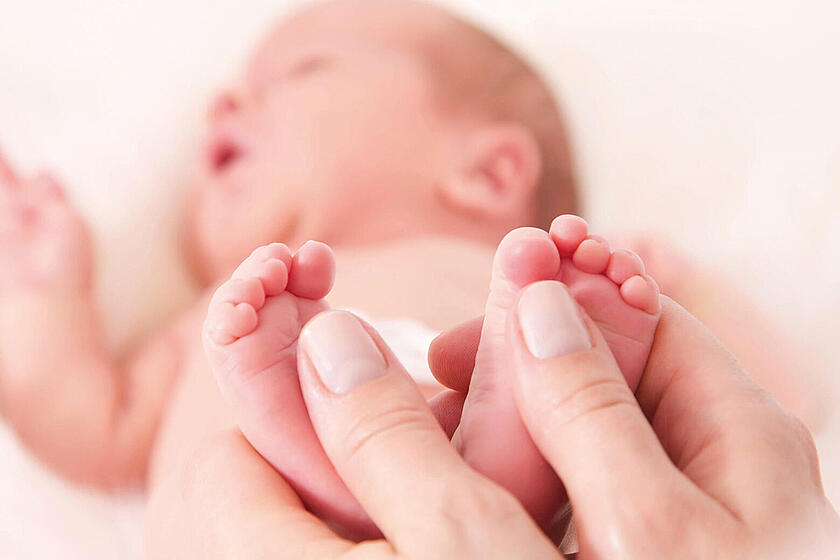 Zwei Hände halten zwei kleine Füße eines Babys. Im Hintergrund sieht man verschwommen das Baby liegen.