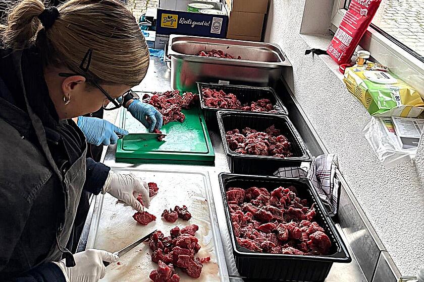 Zwei angehende Feldköchinnen schneiden mit Handschuhen rohes Fleisch auf Schneidebrettern in einer mobilen Küche. Im Hintergrund sind weitere Mitglieder der Johanniter im Einsatz sowie ein Johanniter-Einsatzfahrzeug und Küchenzubehör zu sehen.